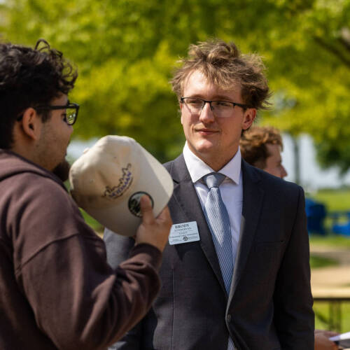 Brody Zondervan, right, in a suit jacket stands outside talking with student in brown hoodie who is holding a tan hat